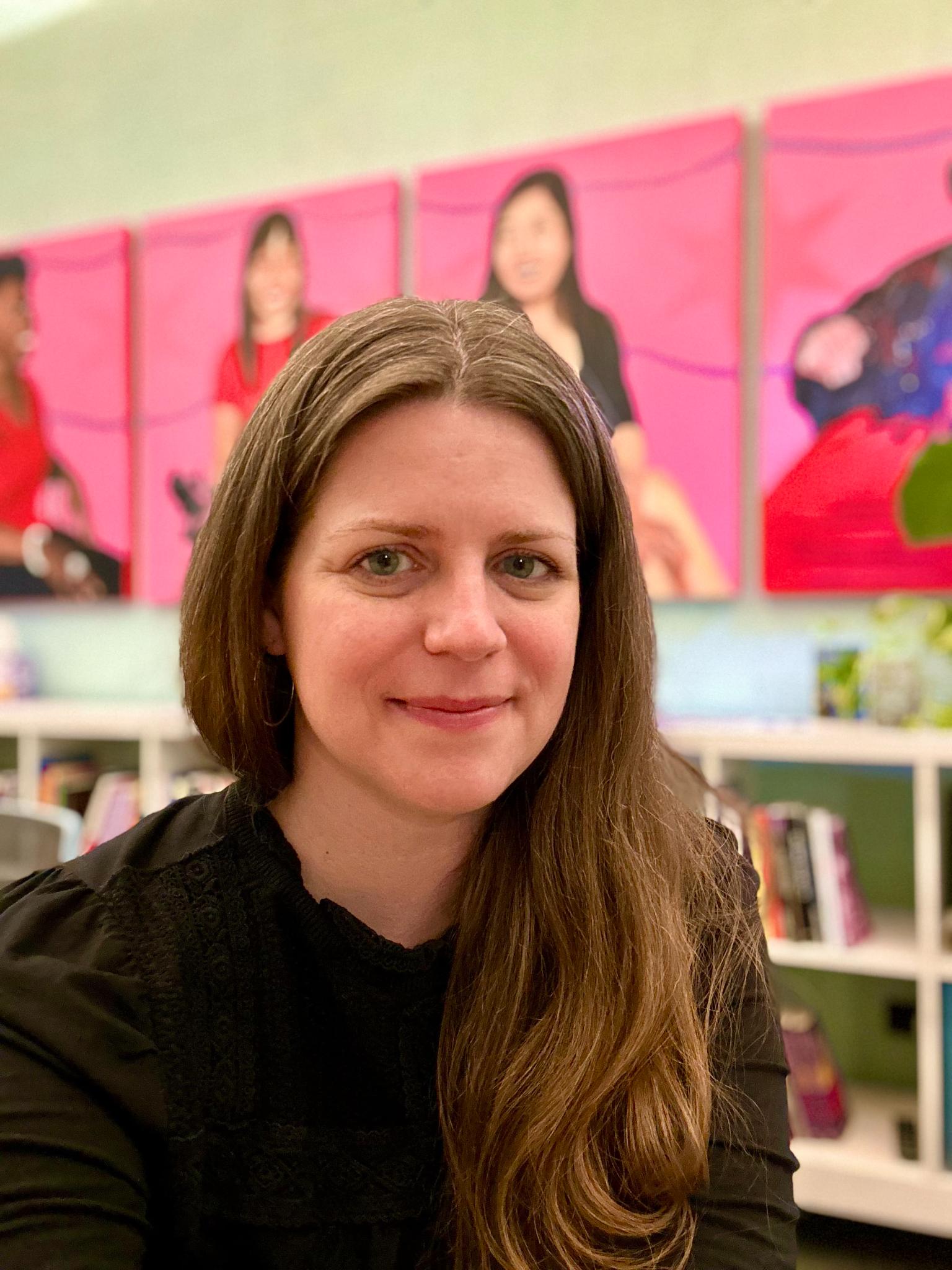  A photo of Margaret Fink, a white woman with long light brown and gray hair and a black top. She is smiling and sits in front of a blurred background with low white bookcases and a four pink and red paintings of Chicago area disabled women of color, a portrait series called Crip Paint Power by Genevieve Ramos.