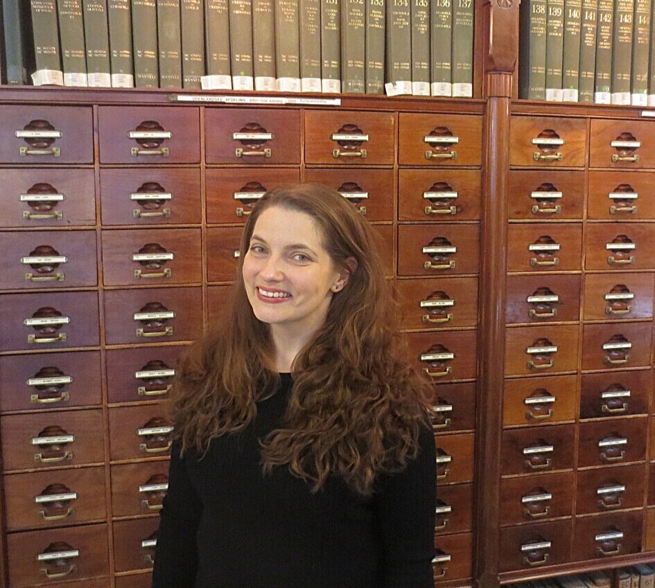 Maura Seale - white woman with long curly hair standing in front of a wooden card catalog